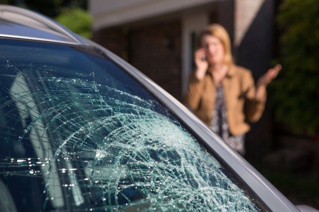A car windshield shattered in a spiderweb pattern, with the owner on the phone in the background.
